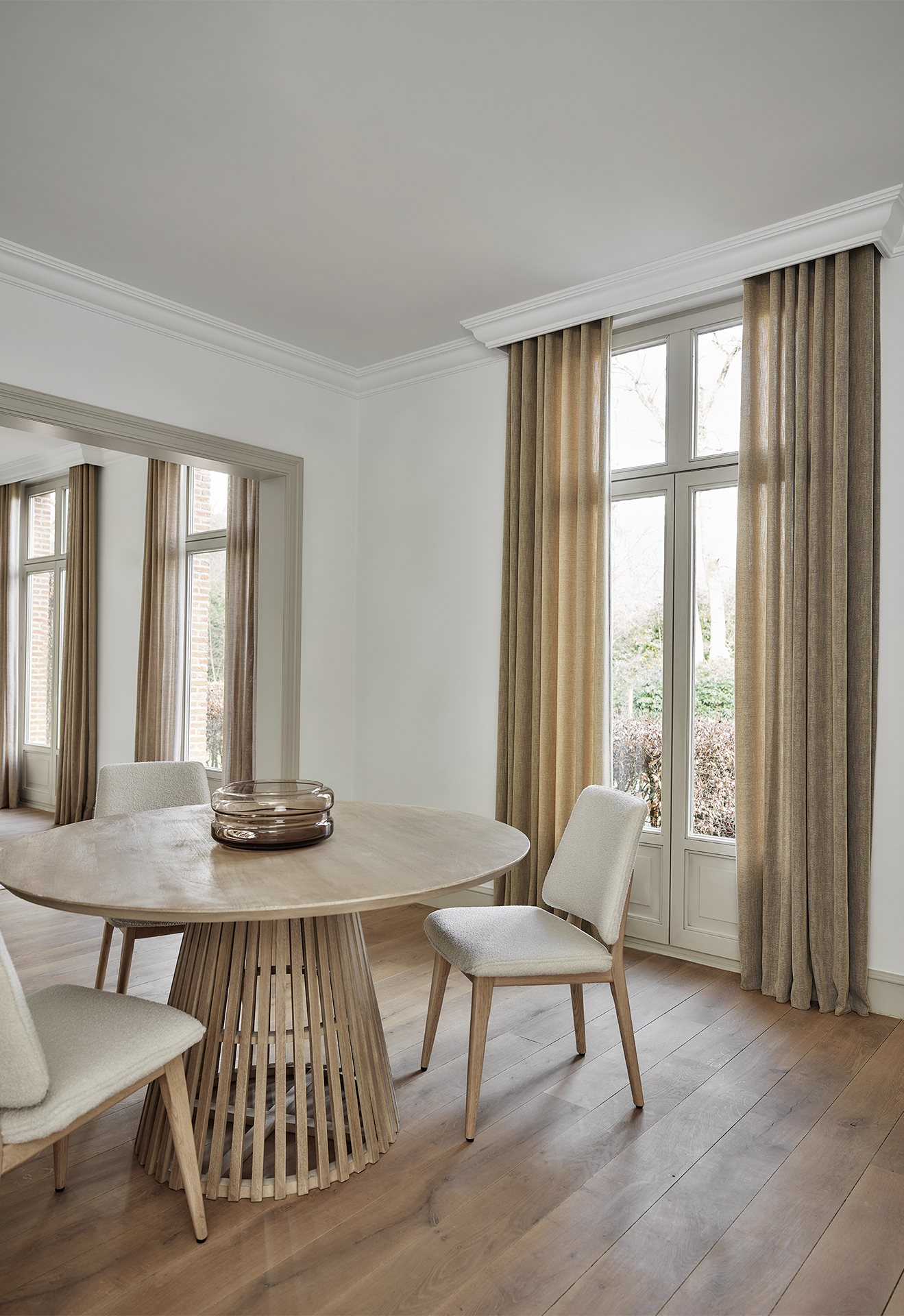 Stue med høye vinduer kledd i gardiner som går helt til gulvet, ved siden av et rundt spisebord i tre med stoler