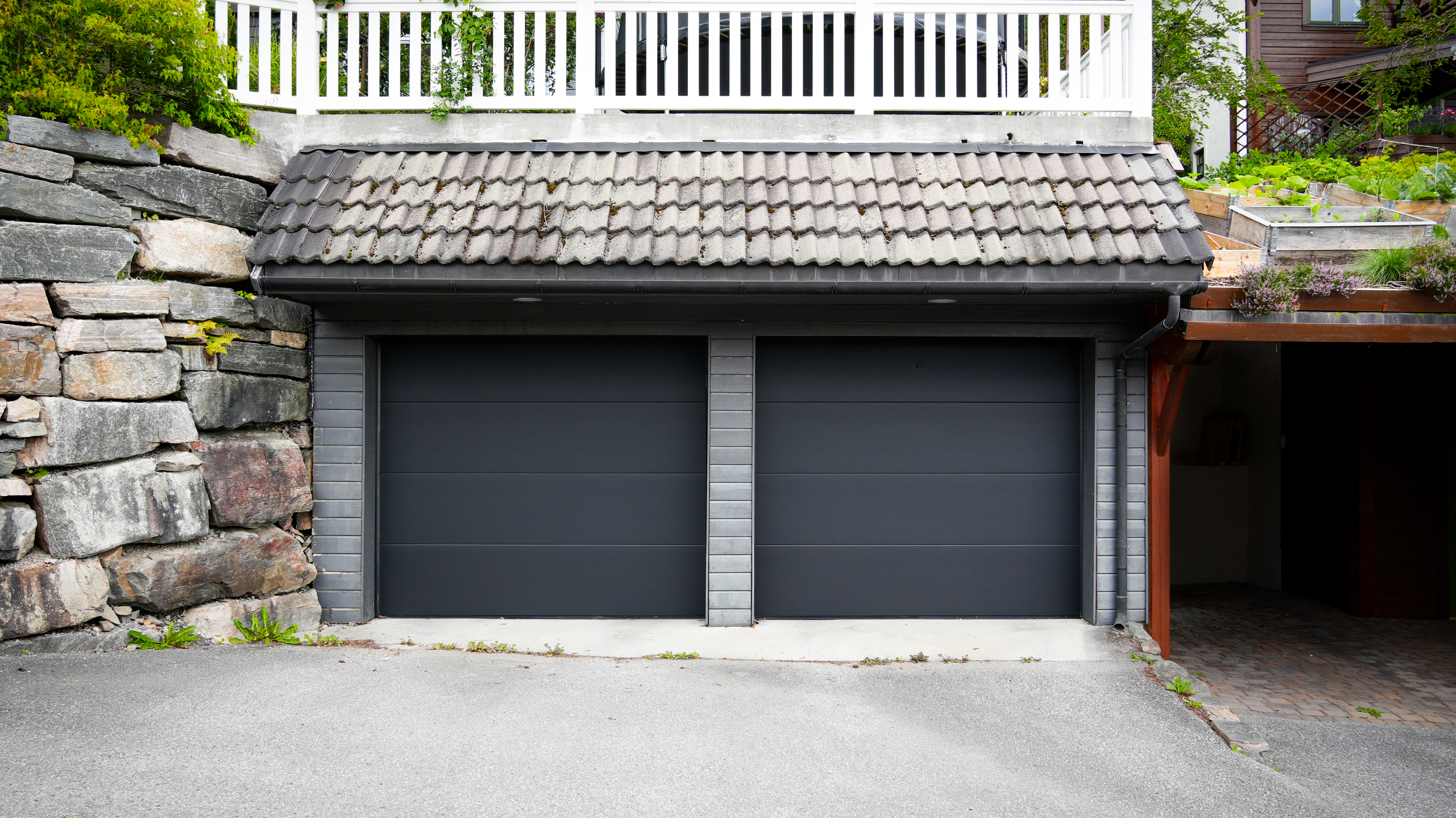 En elegant, grå garasjeport er lukket under en moderne betongstruktur, omgitt av grøntområder og tilstøtende mørke trepaneler.