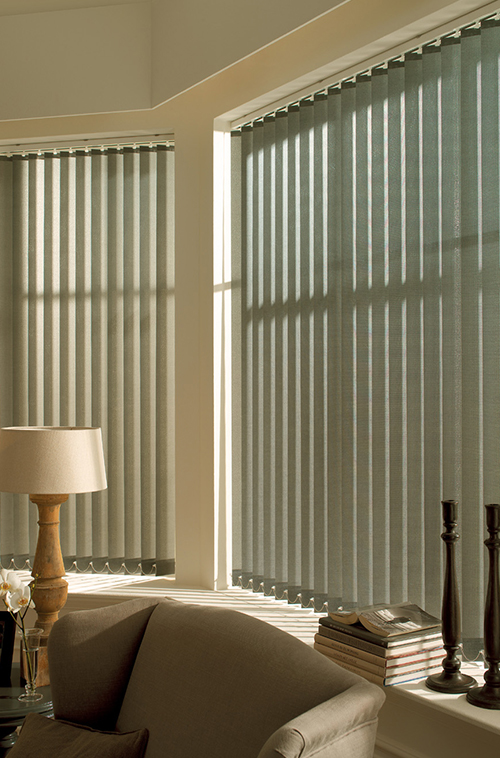 Vertikale persienner delvis åpne, kaster skygger i et lyst rom med en lampe, sofa, stabler med bøker og dekorative gjenstander i nærheten.