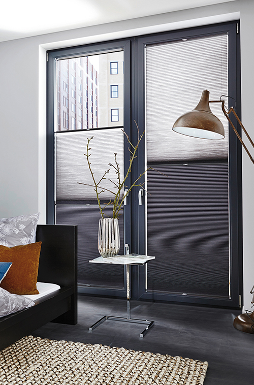 Moderne soverom med store vinduer, pliségardiner, et sidebord i glass og en høy gulvlampe.