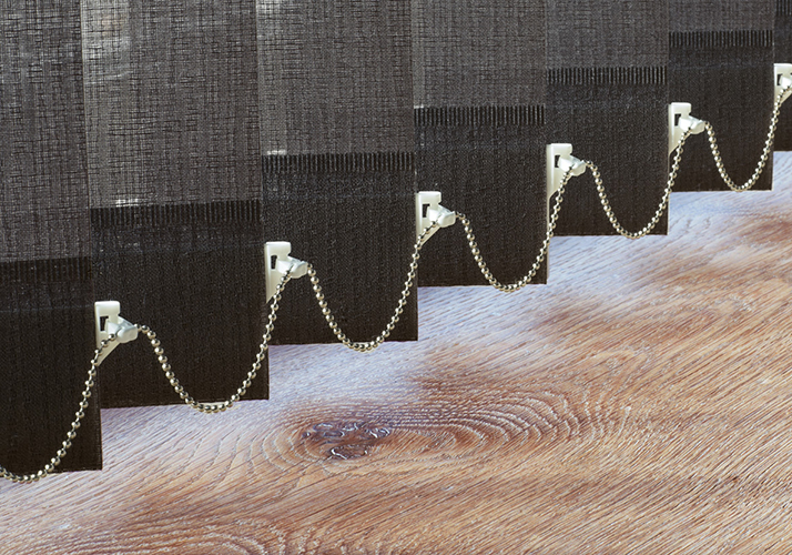 Nærbilde av mørke vertikale persienner med lenket kulekjede nederst over et tregulv.