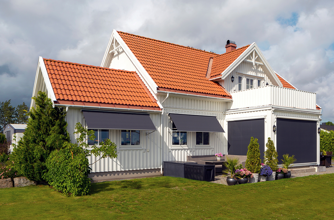 Hvitt hus med rød tegltak og svarte, uttrekkbare markiser over vinduer og uteområde.