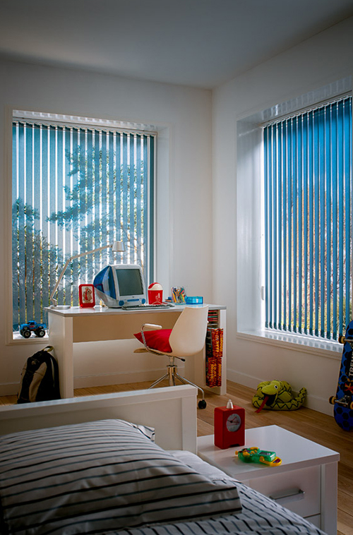 Lyst soverom med stripete sengetøy, pult ved vinduene med blå vertikale persienner og spredte leker.