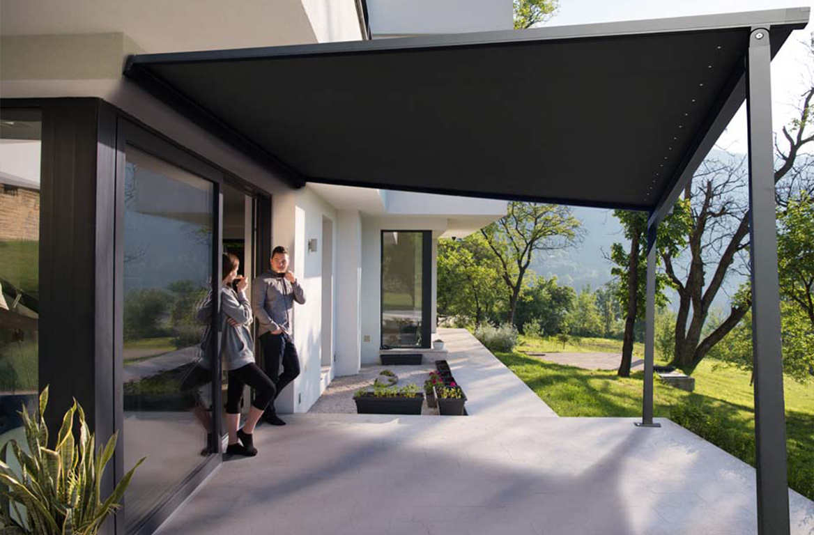 Moderne terrasse med svart markise, store glassdører og to personer som prater med grønn natur som bakgrunn.