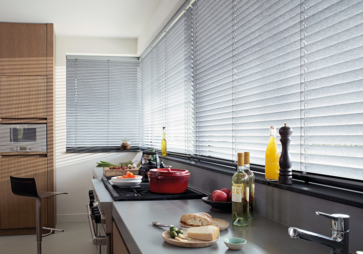 Lys, moderne kjøkken med lang benkeplate, matforberedelser og store vinduer med elegante grå persienner.