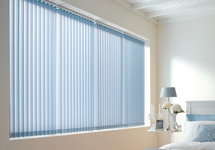 Lyst soverom med stort vindu dekket av lyseblå vertikale persienner, hvite møbler og myke interiørdetaljer.