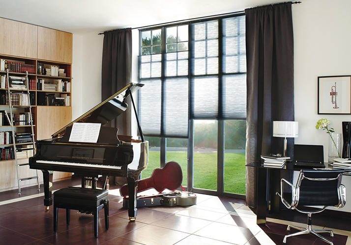 Elegant musikkrom med flygel, bokhyller, skrivepult og høye vinduer med grå pliségardiner og gardiner.