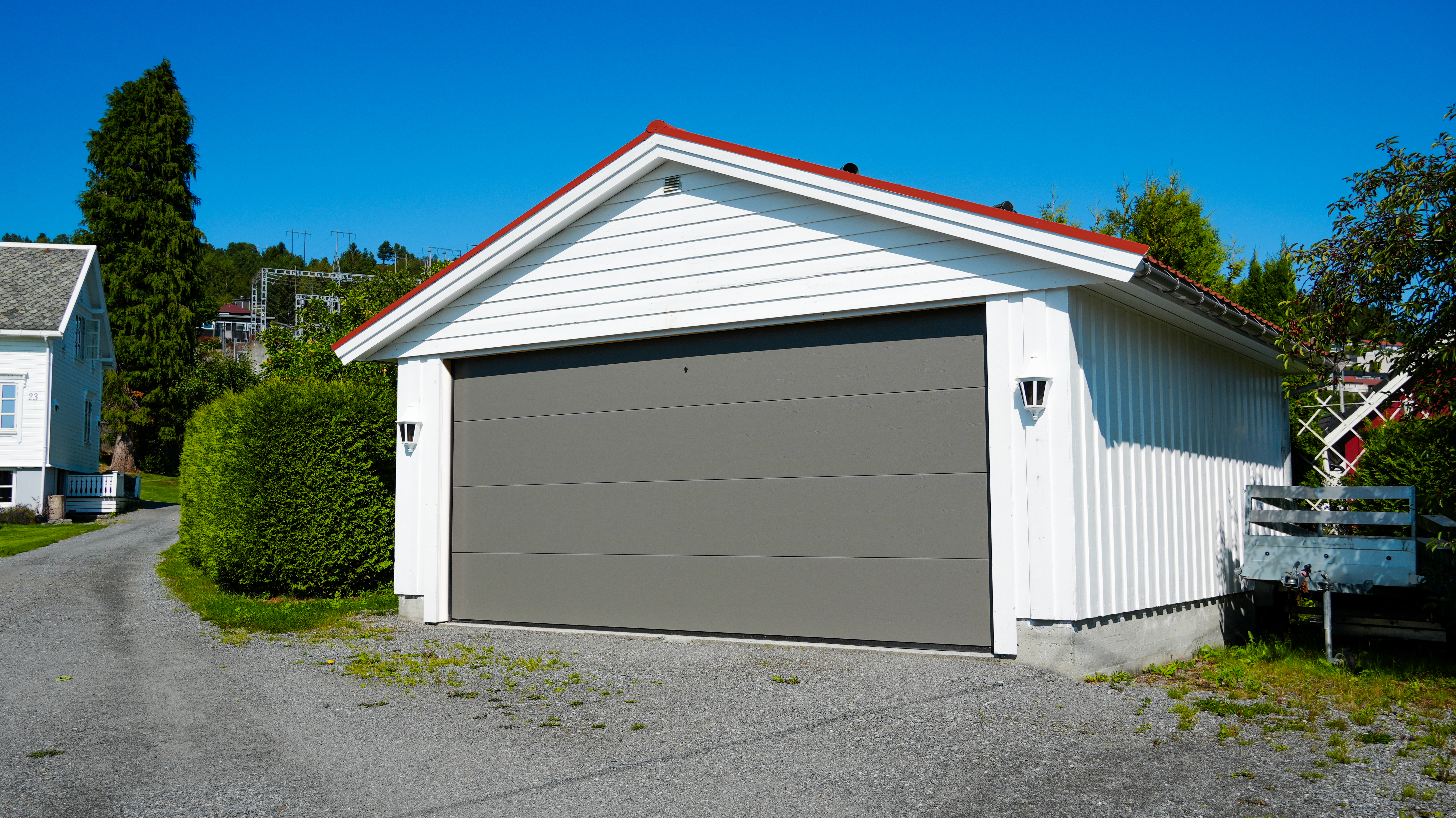 En hvit garasje med lukket grå port står ved siden av en gruslagt oppkjørsel, omgitt av grønt og klar blå himmel.