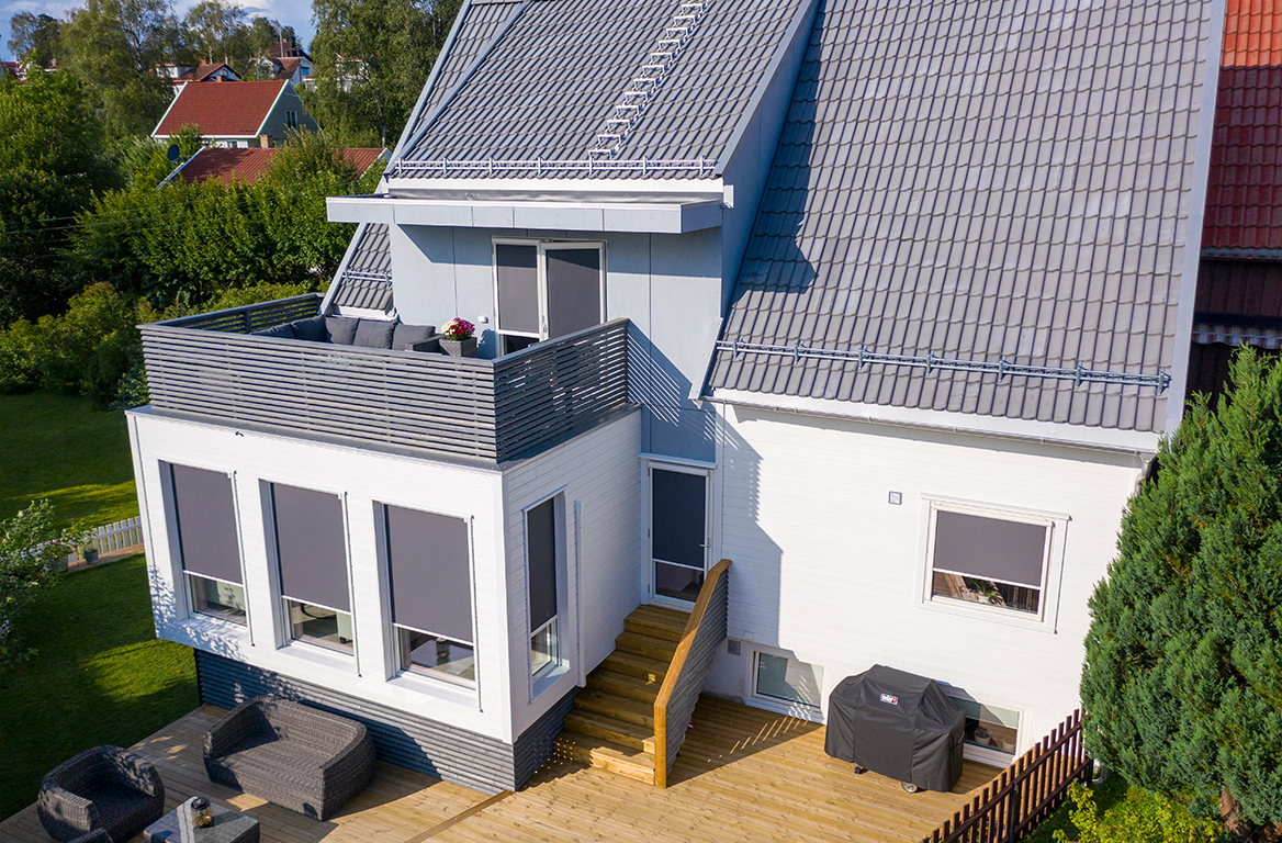 Moderne hvitt hus med grått tak, sitteplasser på balkongen og store vinduer utstyrt med elegante grå utvendige persienner.