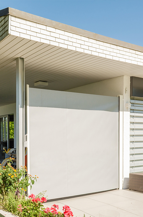 Hvit, moderne terrasse med vertikal avskjermingsvegg, hageblomster og flatt tak under klar blå himmel.