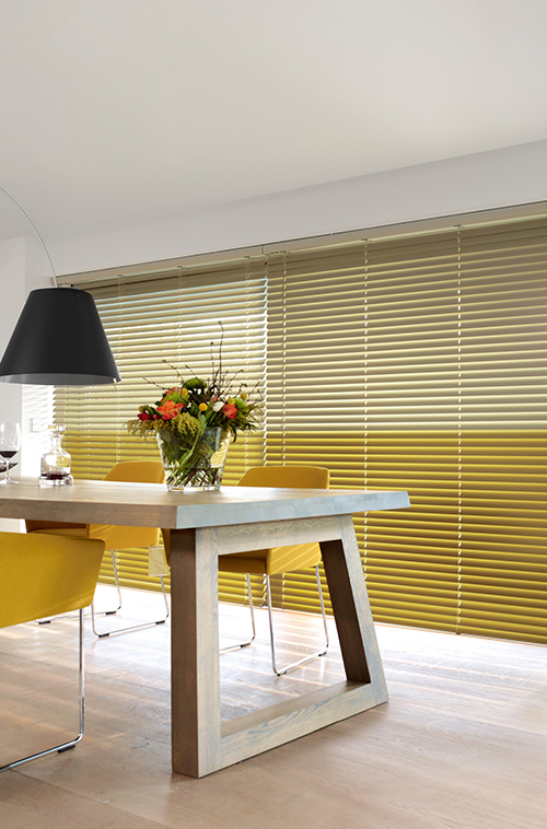Spisestue med trebord, gule stoler, sort pendellampe og bredt vindu dekket av gule persienner.