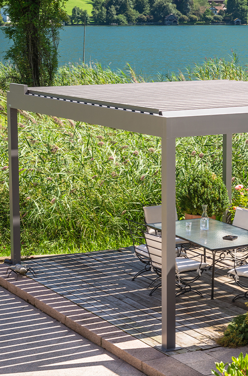 Uteplass med moderne pergola, glassbord og stoler med utsikt over en innsjø omgitt av grønt.