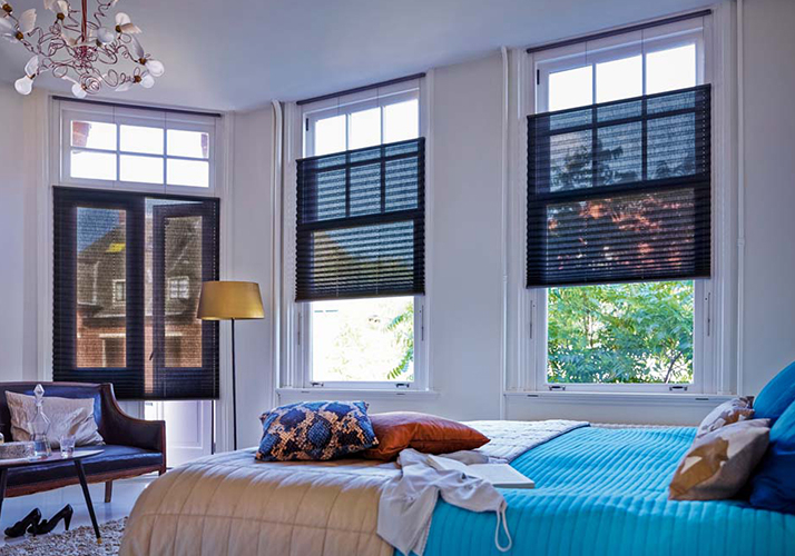 Koselig soverom med blått sengetøy, lenestol og høye vinduer med mørke plisségardiner.