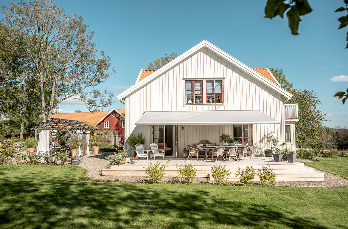 Hvitt trehus med rødt tak og stor terrasse, skygget av en bred uttrekkbar markise i hagen.