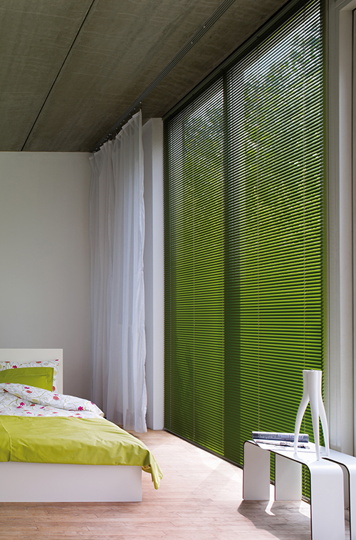 Minimalistisk soverom med grønt sengetøy, lette gardiner og høye vinduer med grønne persienner.