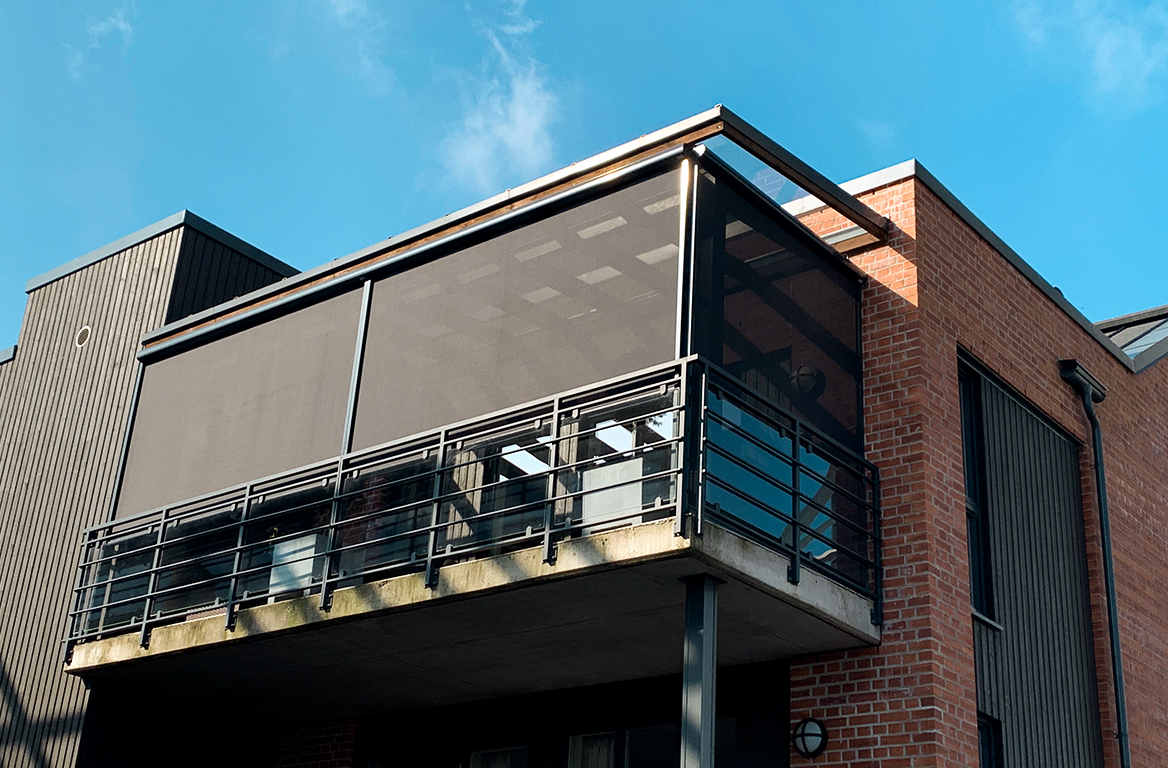 Moderne murbygning med balkong omsluttet av grå netting og svarte rekkverk under blå himmel.
