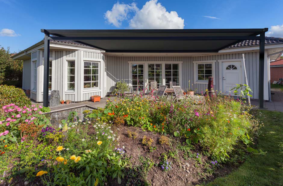 Hvitt hus med svart terrassemarkise, utendørs spiseplass og fargerik blomsterhage foran.