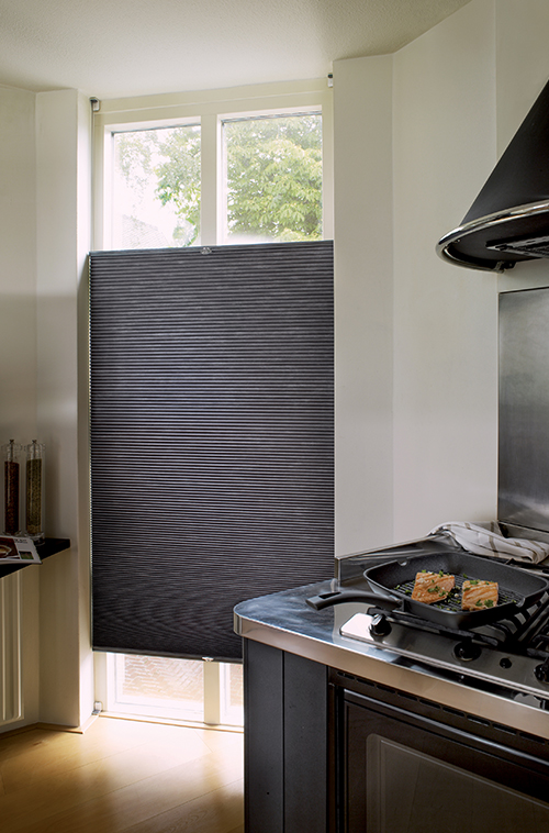 Kjøkkenkrok med svart plisségardin på høyt vindu, rustfrie hvitevarer og laks som grilles på komfyren.