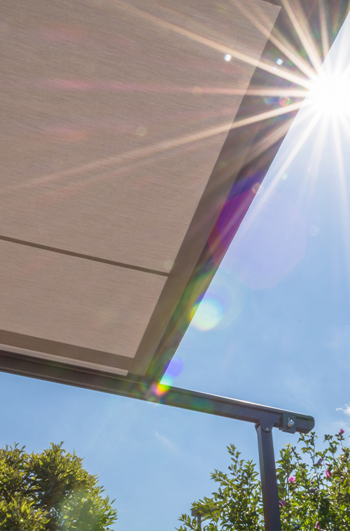 Beige uttrekkbar markise sett nedenfra, med sollys og linsebluss mot blå himmel og trekroner.