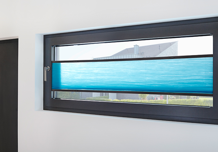 Bredt horisontalt vindu med knallblå plissert gardin delvis trukket ned, innrammet i mørkt treverk med utsikt mot hagen.