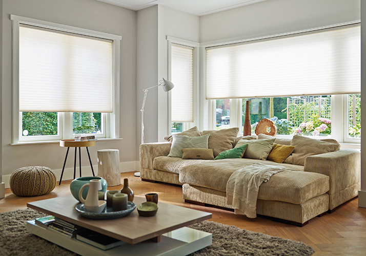 Koselig stue med beige hjørnesofa og store vinduer dekket med hvite gardiner.