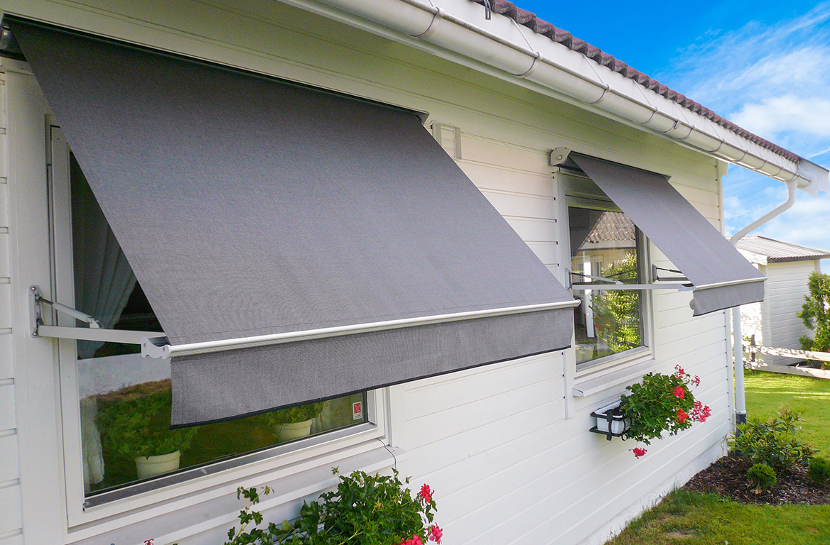 Hvit husfasade med grå markiser trukket ut over to vinduer, og blomsterkasser under.