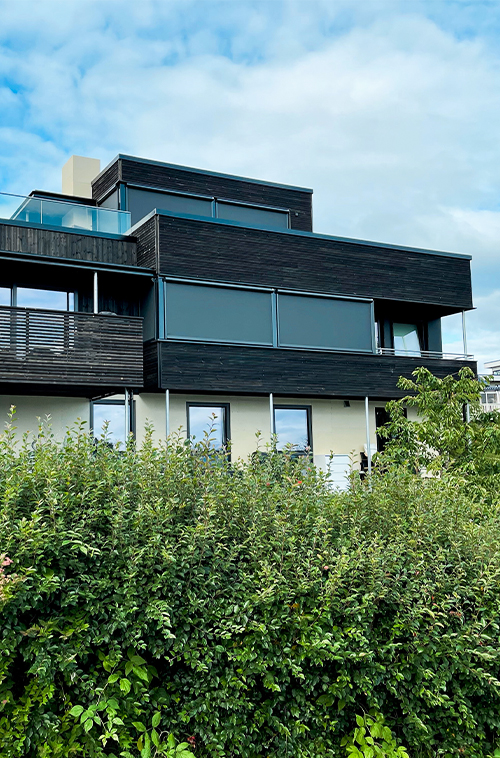 Et moderne hus over flere plan med store glassvinduer, omgitt av frodig grønt landskap under blå himmel.