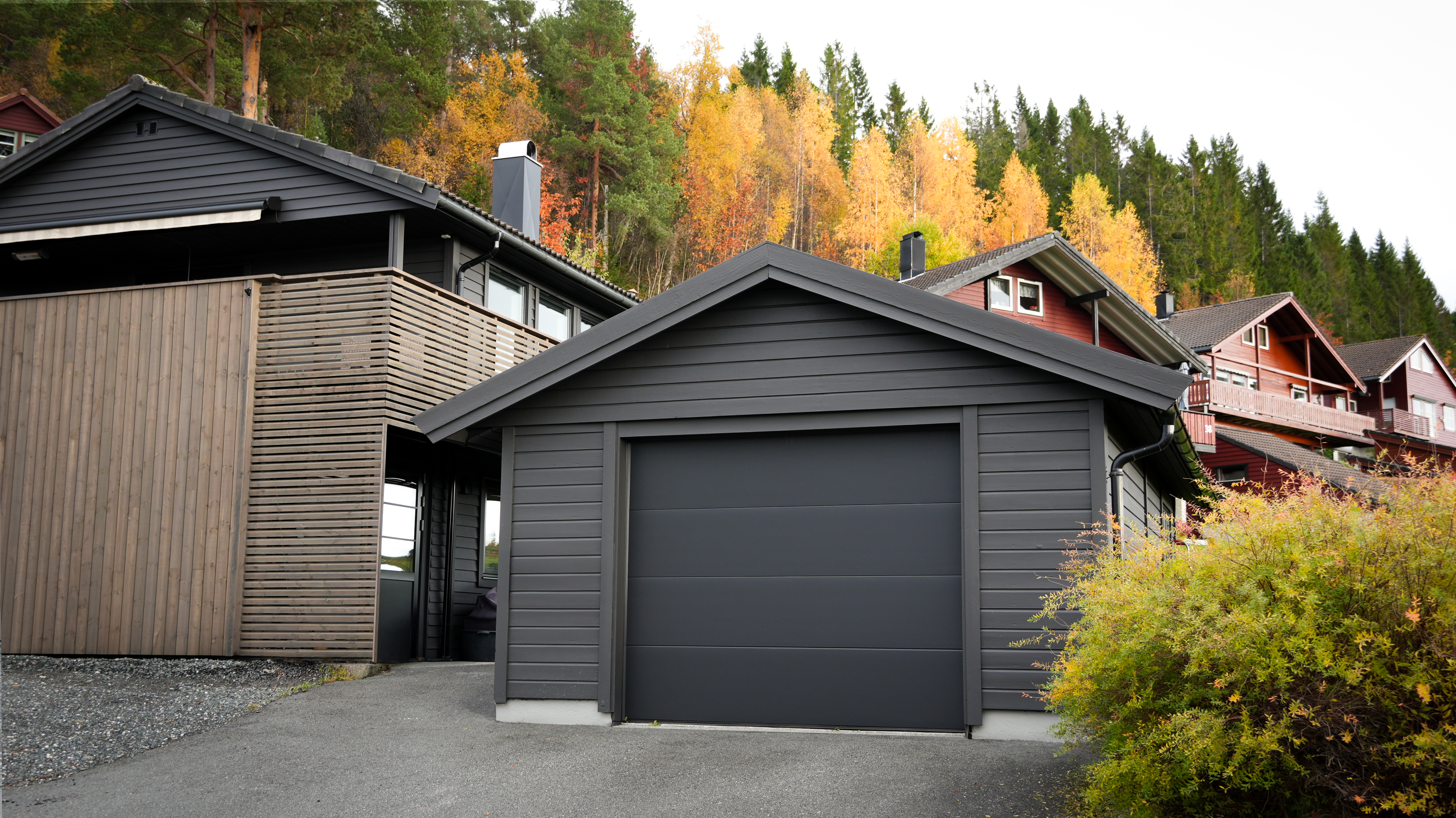 Moderne mørkgrått hus med integrert garasje, omgitt av trær i høstfarger.