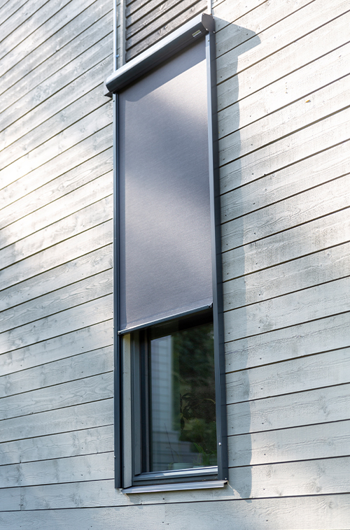Høyt, smalt vindu på en yttervegg i treverk med en elegant grå utvendig rullegardin delvis nedsenket.