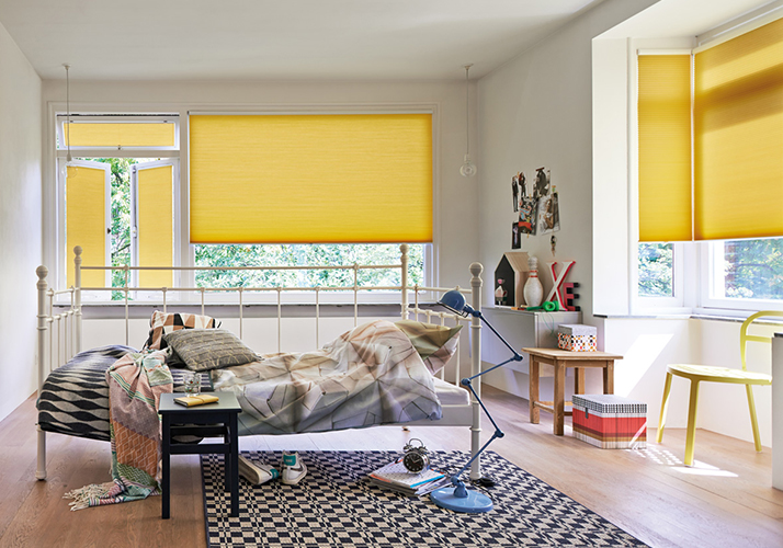 Lys soverom med hvite vegger, tregulv og gule gardiner som gir varme til de store vinduene.