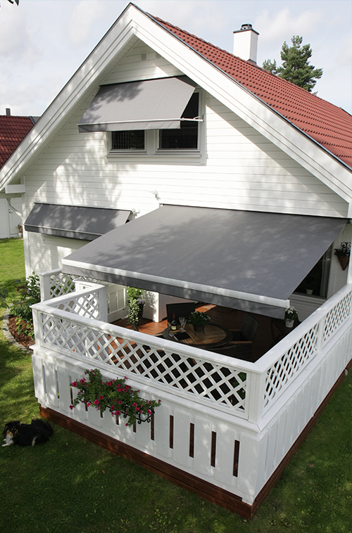 Hvit bolig med rødt tak og grå uttrekkbare markiser over vinduene samt en overbygd veranda med espalierrekkverk.