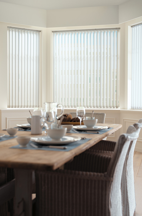Spisestue med rottingstoler rundt et trebord, med høye vinduer og hvite vertikale gardiner.