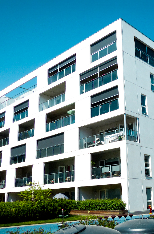 Moderne hvitt leilighetsbygg med flere balkonger, hver utstyrt med mørke uttrekkbare rullegardiner.