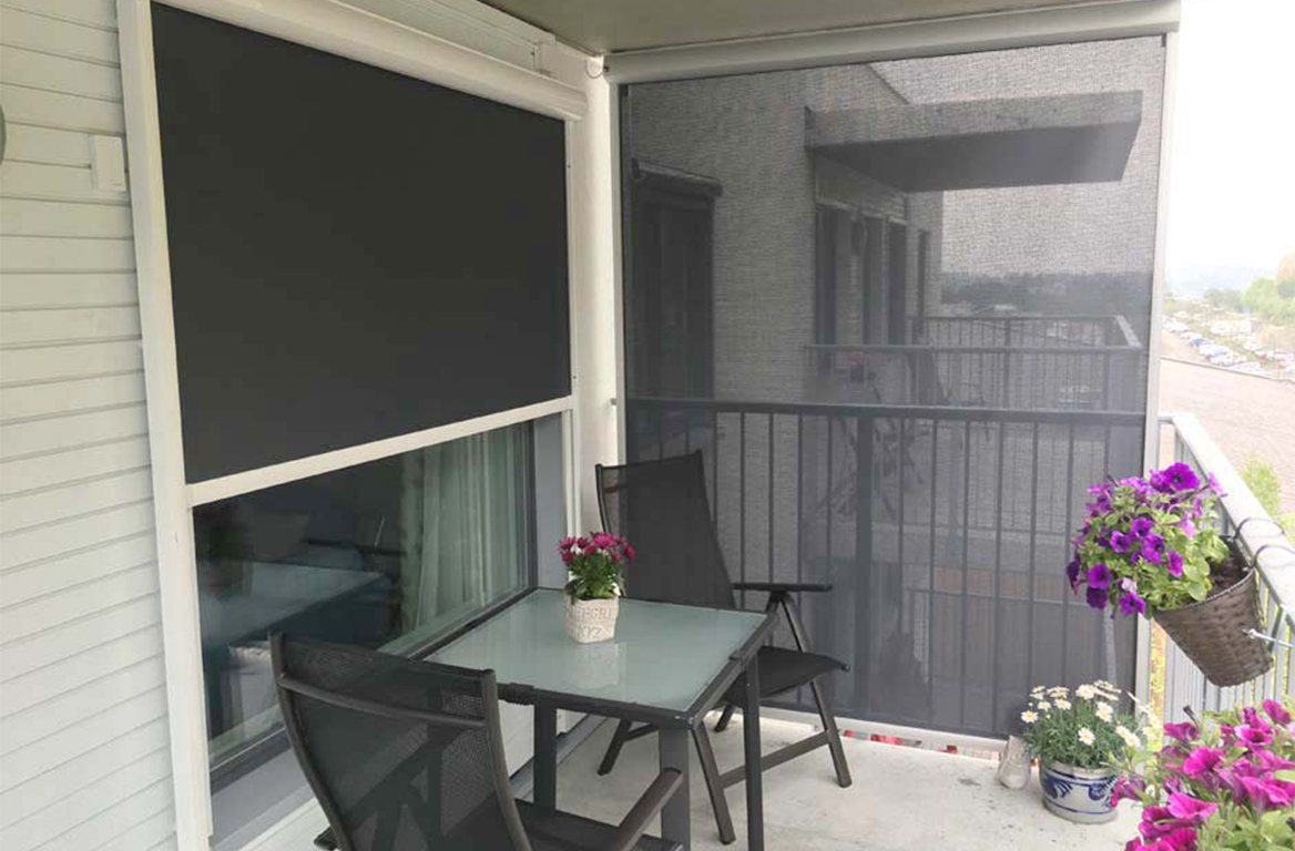 Liten balkong med svarte utvendige rullegardiner, glassbord, stoler og blomsterpotter som skaper en koselig krok.