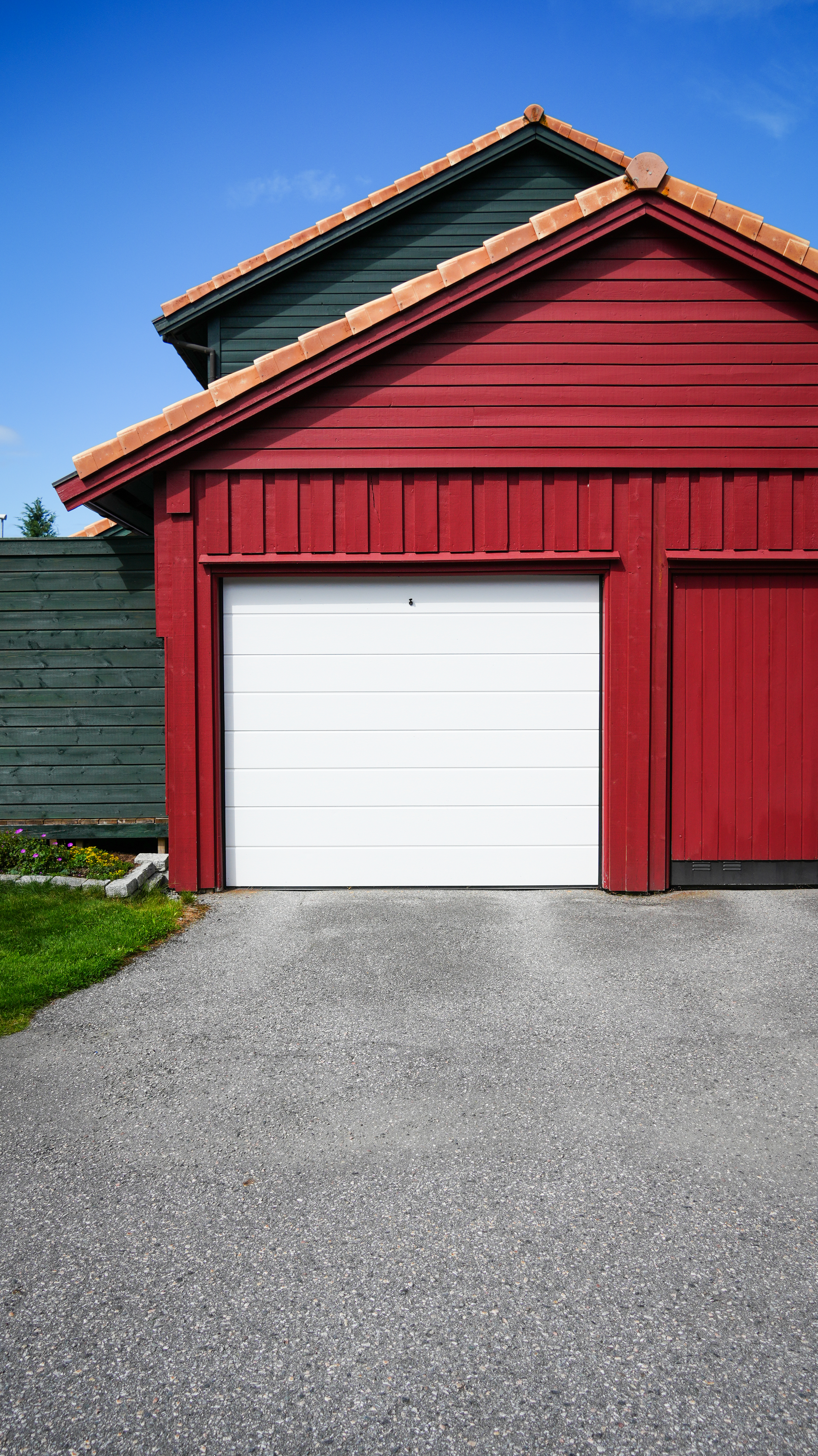 En rød garasje med lukket hvit garasjeport, omkranset av grønt gress og gjerde, under en klar blå himmel.