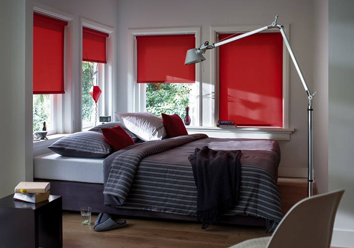 En grå seng med puter står foran store rødfargede vinduer som gir et varmt lys, i en moderne stue.