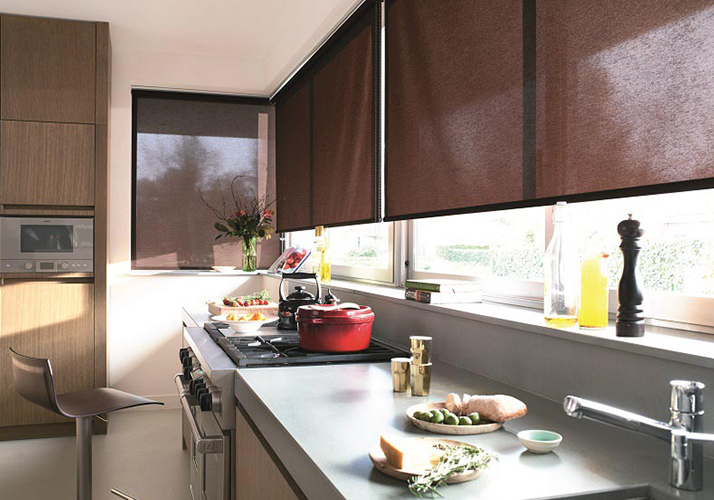 Moderne kjøkken med brune rullegardiner over store vinduer, slanke benkeplater og matlagingsområde.