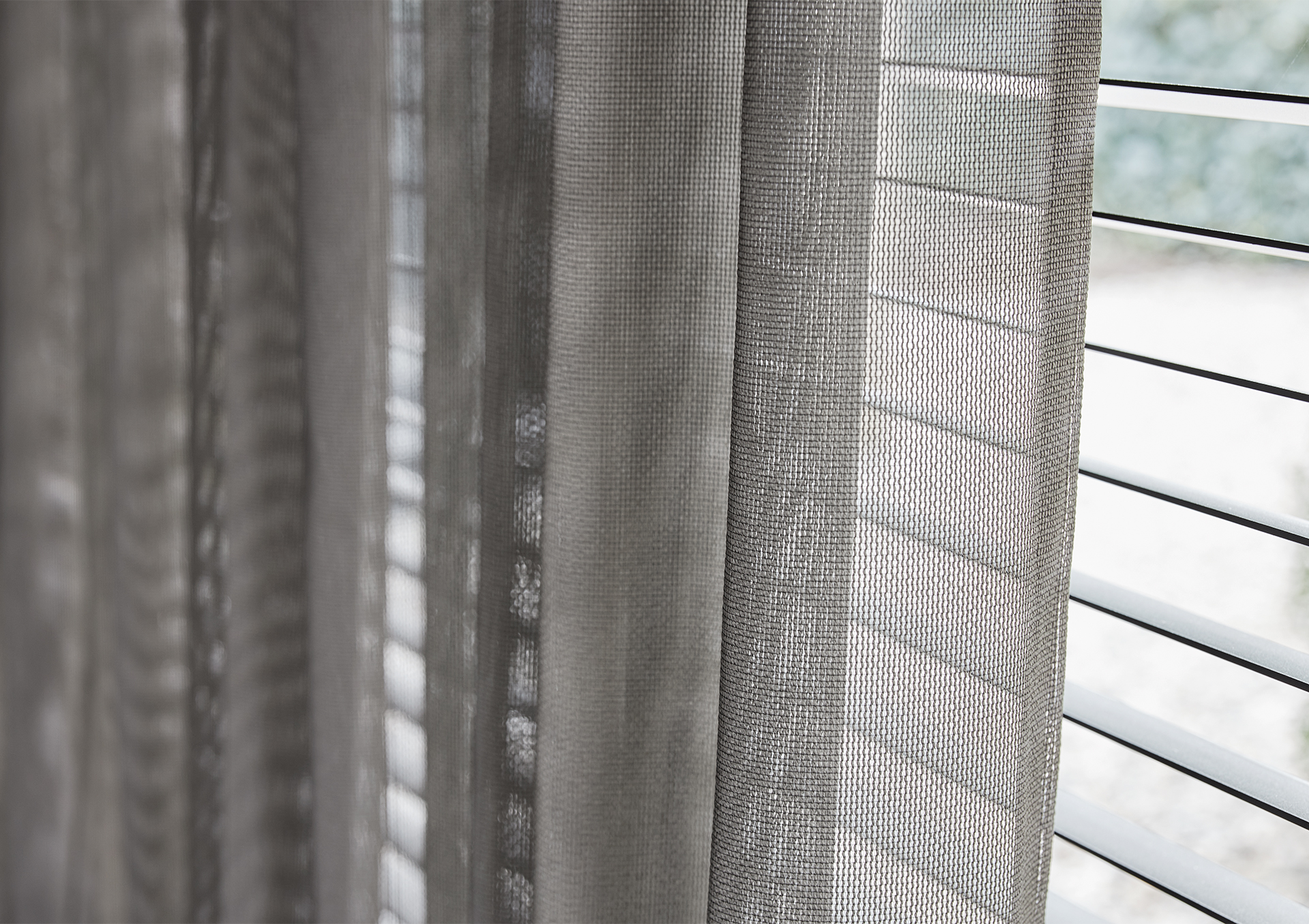 Nærbilde av gjennomsiktige grå gardiner kombinert med horisontale persienner, som mykt filtrerer dagslyset inn i rommet.
